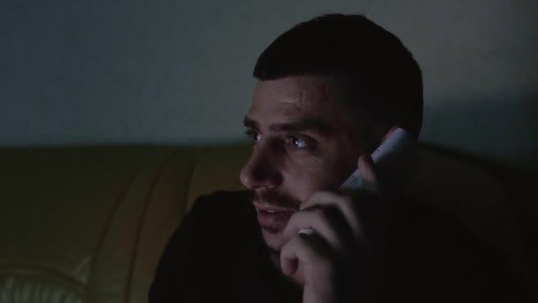 Мужчина разговаривает по беспроводному радио-телефону, смеется и улыбается, сидя по ночам на диване один дома и смотрит видео по телевизору в квартире. Молодой 30-летний кавказский мужчина — стоковое видео