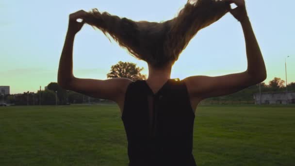 Flickan kastar sitt långa lockiga hår med händerna vid solnedgången i slow motion. Frihetskoncept, nöje, semester, vila. Bakifrån på baksidan. Parkera utomhus. Närbild. Steadicam skjuten — Stockvideo