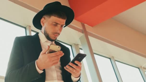 Homem vestindo terno preto e chapéu com dois smartphones decide seus próprios assuntos. A pessoa parece um judeu ou uma máfia italiana. Tiro de ângulo baixo, vista de baixo. Empresário com barba e bigode — Vídeo de Stock