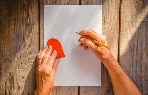 Elderly woman hand going to write a letter, red heart on rustic wooden table as a background, sun rays. View from above, copy space. Mother or Woman, Valentine day, human emotions, love and compassion, warmth, senior concept.