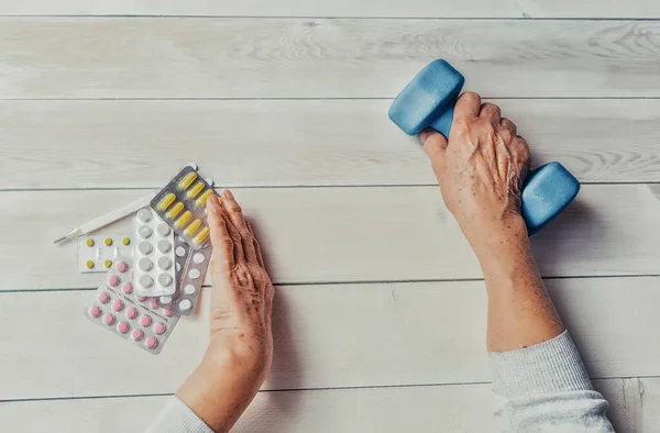 Senior hands with pills, drugs on table, dumb-bell. Woman wrinkled hands, thermometer, colorful tablets and dumb-bell, wooden background. Health care for the elderly, healthy living concept.