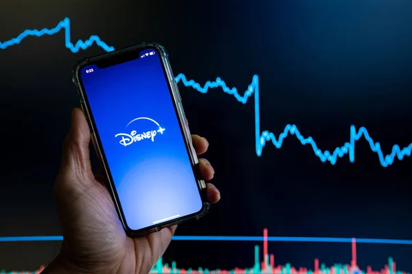 Disney Plus Disney app held against a dark stock trading chart going down in value — Stock Photo, Image