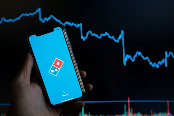 Dominos pizza mobile delivery app held against a dark stock trading chart going down in value — Stock Photo, Image