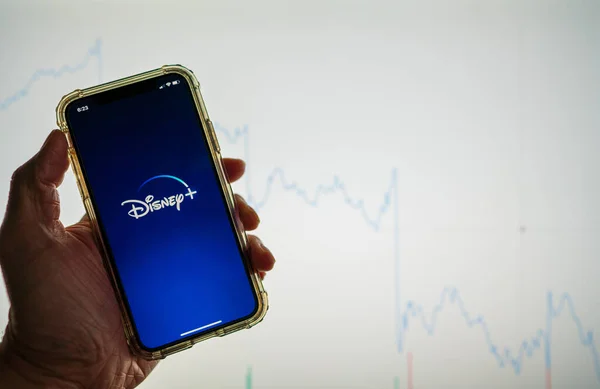 Disney Plus Disney mobile app held against a white stock market chart going down in value — Stock Photo, Image