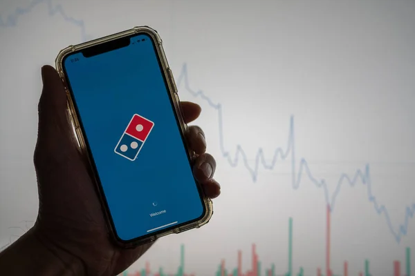 Domino s pizza mobile app held against a white stock trading chart going down in value — Stock Photo, Image