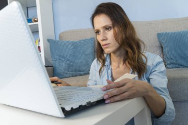 Genç bir kadın, beyaz bir masada dizüstü bilgisayarı olan bir evden bilgisayar için çalışıyor.