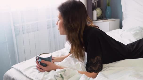 黒いバスローブを着たサイドビューの女性がベッドに横になり コーヒーを飲みながら朝のコーヒーを楽しんでいます — ストック動画