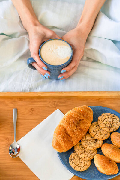 Breakfast in bed with fruits and pastries on a tray and womans hands hold cup of coffee. Vertical photo