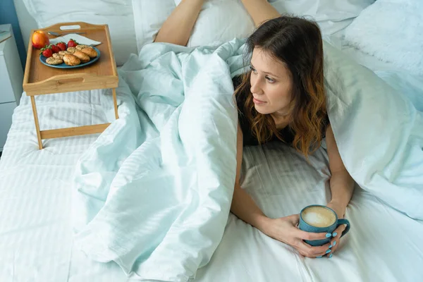 Young funny woman having a breakfast on tray on the bed with cup of coffee under coverlet. Happy morning on vacation