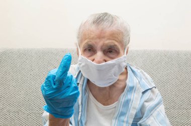 Tıbbi maskeli yaşlı bir kadın ve orta parmağını gösteren bir el kaldırmış. Covid-19 virüs salgını ve dünya çapında salgınla savaşmak için orta parmağını gösteriyor. Coronavirus reddetme konsepti
