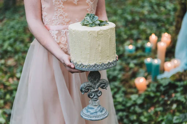 Bride in blush wedding dress holding an unusual wedding cake decorated with succulent plant with forest floor in the background