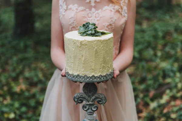 Bride in blush wedding dress holding an unusual wedding cake decorated with succulent plant with forest floor in the background