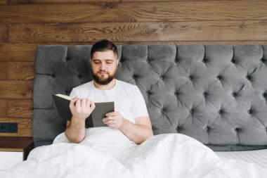 Young handsome caucasian man reading a book sitting in bed