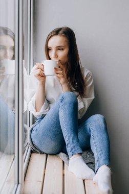 Yatağında bir fincan kahve olan çekici bir kadın. Pencerenin önünde oturup çay içen güzel kız. Bir bardak sıcak içeceği olan kadın..