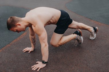 Çıplak gövdeli ve siyah şortlu genç bir adam spor sahasının kenarındaki barda duruyor.