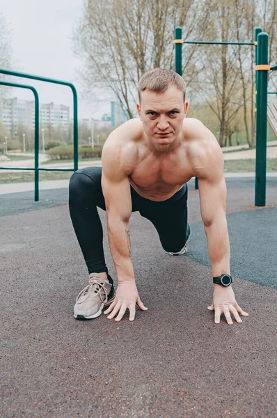 A young guy stretches and warms up on the sports ground. Muscular sports guy doing fitness outdoors.