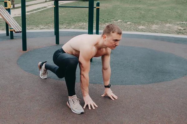 A young guy stretches and warms up on the sports ground. Muscular sports guy doing fitness outdoors.