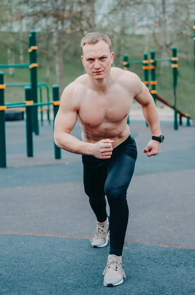 A young guy stretches and warms up on the sports ground. Muscular sports guy doing fitness outdoors.