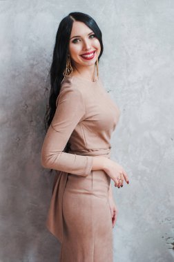 İçerideki dokulu bir duvarın arka planında bej bir elbiseyle model görünümlü mutlu siyah saçlı kız.