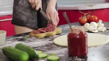 Genç kız pizza pişirir ve ahşap kesme tahtasıyla füme sosis keser, kör bıçakla.