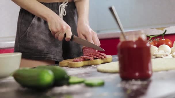 Chica joven cocinar una pizza y cortar una salchicha ahumada en una tabla de cortar de madera, cuchillo opaco, mesa negra — Vídeos de Stock