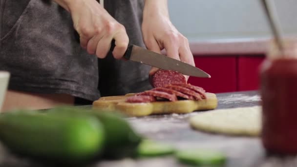 Jeune fille cuisiner une pizza et couper une saucisse fumée sur une planche à découper en bois, couteau terne, table noire — Video