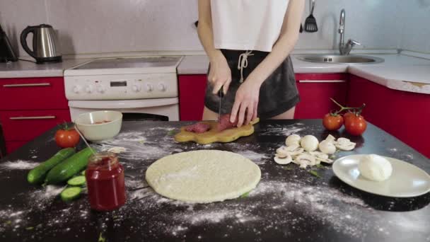 Młoda dziewczyna gotować pizzę i wyciąć wędzoną kiełbasę na drewnianej desce do krojenia, tępy nóż, czarny stół — Wideo stockowe