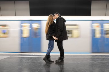 Genç çift metroda hareket halindeki bir trenin arka planında öpüşüyor.