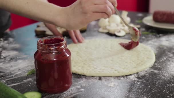 Χέρια ενός νεαρού κοριτσιού πασπαλίζουν πελτέ ντομάτας σε ζύμη πίτσας — Αρχείο Βίντεο