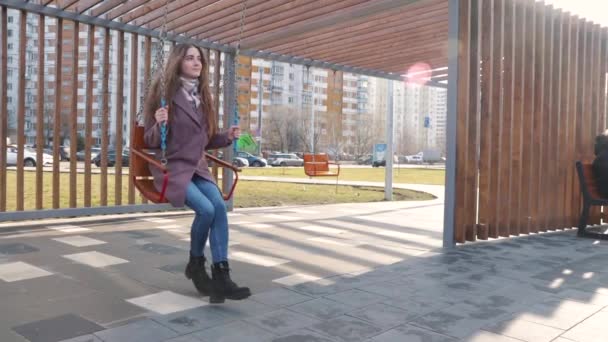 Gelukkig jong meisje swinging op een swing in het park met auto 's in de achtergrond — Stockvideo