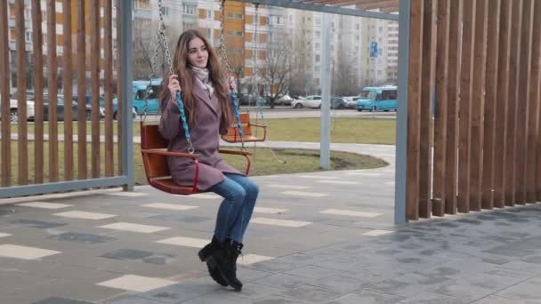 Feliz chica joven balanceándose en un columpio en el parque con coches en el fondo — Vídeos de Stock