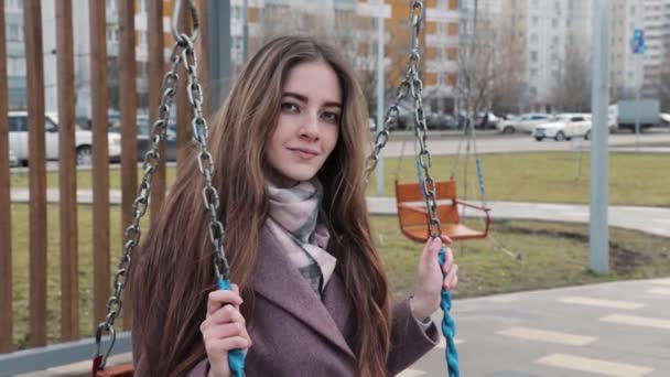 Glad ung flicka svänger på en gunga i parken med bilar i bakgrunden — Stockvideo
