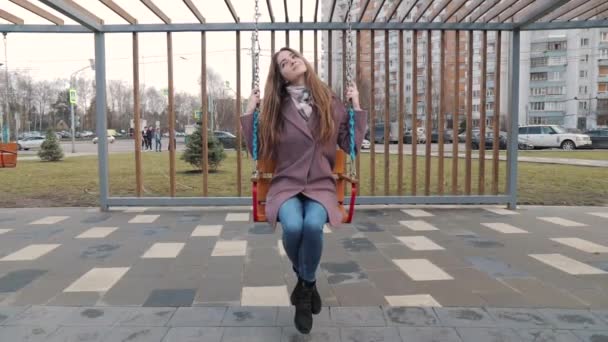 Feliz chica joven balanceándose en un columpio en el parque con coches en el fondo — Vídeos de Stock