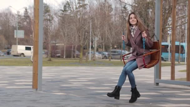 Glad ung flicka svänger på en gunga i parken med bilar i bakgrunden — Stockvideo