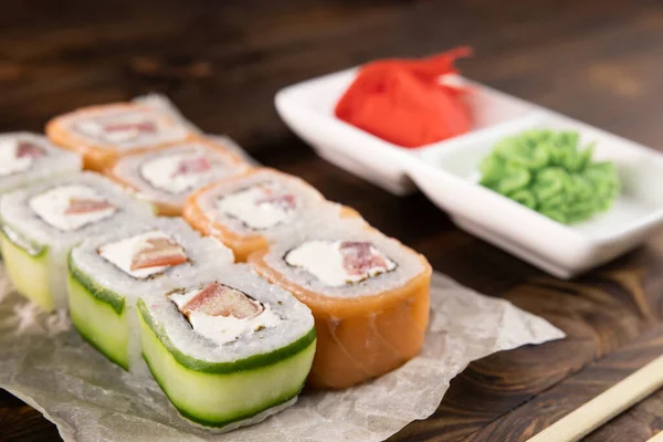 Kağıt üzerinde somon, domates, peynir ve salatalık. Kare tabaklarda Wasabi ve zencefil. Koyu tahta masada.