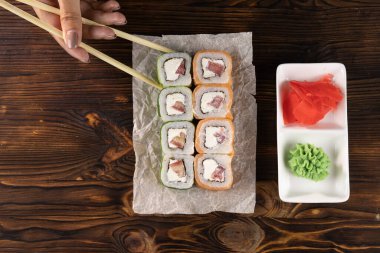 Elleri somonlu, domatesli, peynirli ve salatalıklı rulo alıyor. Wasabi ve zencefil kare tabaklar üzerinde koyu ahşap masa üstünde.