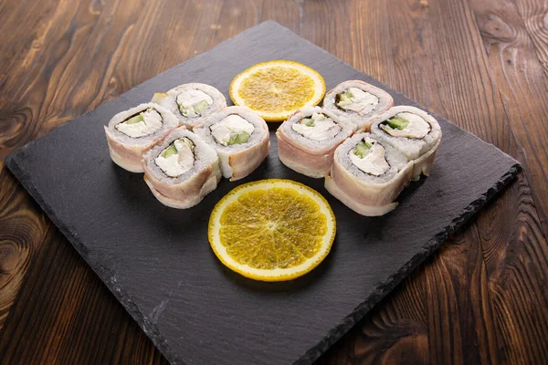 Sushi rolls with bacon, avocado and white fish with sliced orange on the black japan plate