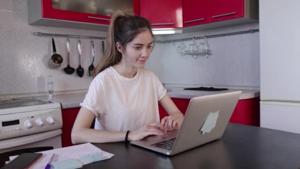 Молодая девушка работает с ноутбуком и разговаривает по телефону на кухне дома — стоковое видео