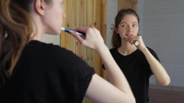 Chica cepilla los dientes en el baño mirando en su reflejo en el espejo — Vídeo de stock