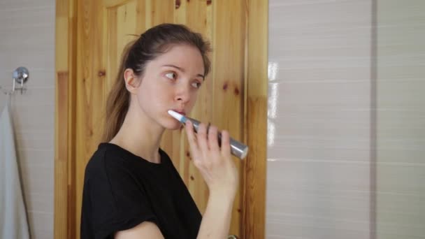 La chica se cepilla los dientes con un cepillo ultrasónico y se mira en el espejo del baño con toalla blanca y puerta de madera en el fondo — Vídeos de Stock