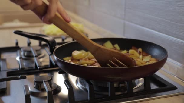 The hand lifts the lid of the pan, stirs the fried potatoes with a wooden spatula on a gas stove — Stock Video