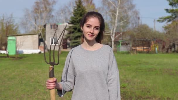 Glückliches junges Bauernmädchen blickt in die Kamera und hält eine Mistgabel mit Fichte und Birke im Hintergrund — Stockvideo