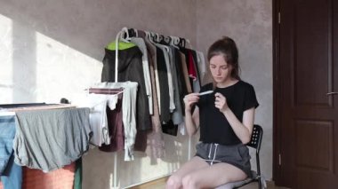 Kadın sandalyeye oturur ve arka planda giysileri olan tırnak törpüsüyle tırnaklarını keser.