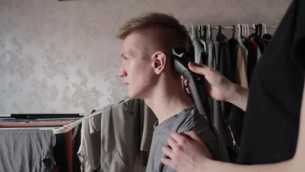 Молодая женщина стрижет мужчину с помощью стрижки волос дома во время изоляции с одеждой на перилах на заднем плане — стоковое видео