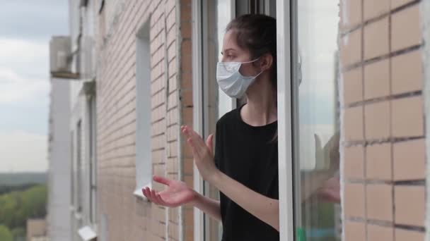 Γυναίκα σε μια ιατρική μάσκα κοιτάζει έξω από ένα ανοιχτό παράθυρο και χειροκροτεί για την υποστήριξη των εργαζομένων στον τομέα της υγειονομικής περίθαλψης — Αρχείο Βίντεο