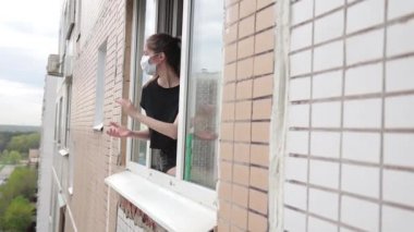 Tıbbi maskeli kadın açık pencereden dışarı bakar ve sağlık çalışanlarını desteklemek için alkışlar. 