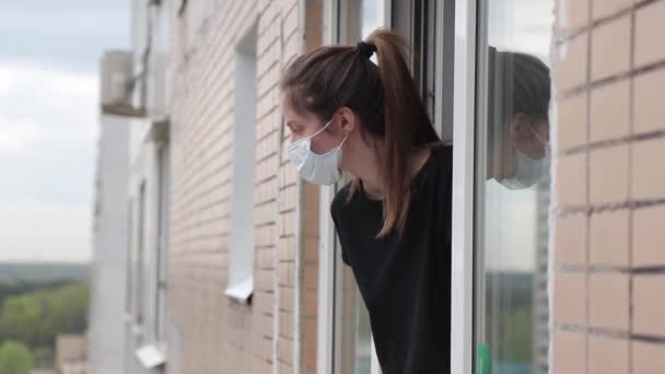 Donna con una maschera medica guarda fuori da una finestra aperta durante l'isolamento — Video Stock