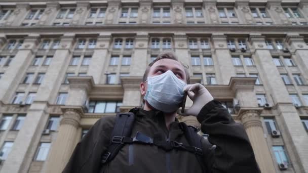 Man met een medisch masker en medische rubberen handschoenen praat aan de telefoon met het gebouw op de achtergrond — Stockvideo