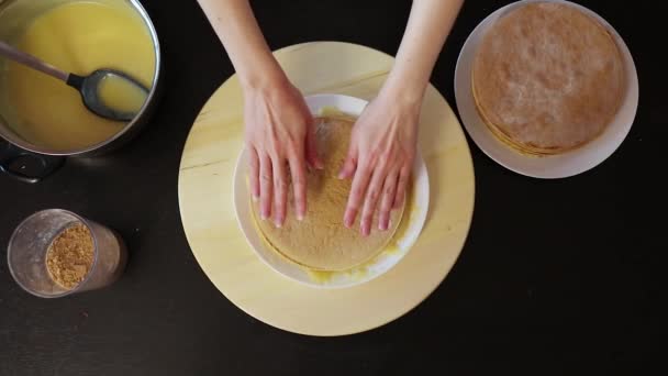 Vrouw neemt een taart, zet het op een ander stuk en smeert crème op de top van een taart die op een ronde houten spinning lade, bovenaanzicht — Stockvideo