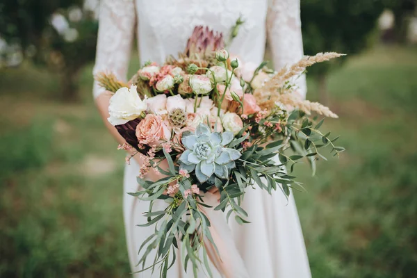 Νύφη σε ένα φόρεμα στέκεται σε ένα καταπράσινο κήπο και κρατώντας ένα γαμήλιο μπουκέτο από λουλούδια και πράσινο — Φωτογραφία Αρχείου